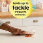 Hand using a cloth to clean up an orange mess on a white surface with text 'holds up to tackle frequent messes' in a yellow circle.